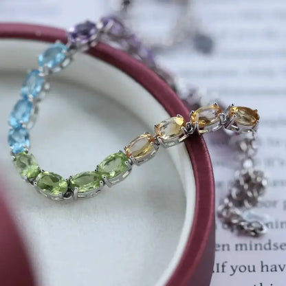 Sterling Silver Natural Crystals Rainbow Tennis Bracelet by Sheehas. The bracelet is made of 925 sterling silver plated with 18K white gold, and is composed of oval-cut garnet, amethyst, topaz, peridot, and citrine arranged in sequence. The bracelet is adjustable in length, suitable for wrists with a circumference of 15cm to 20cm.